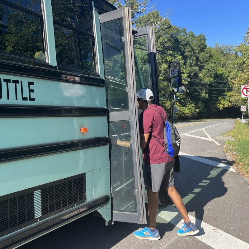 Ảnh người đàn ông lên xe buýt. Anh ấy mặc quần short, áo sơ mi ngắn tay và đeo ba lô. Bầu trời xanh và cây cối.